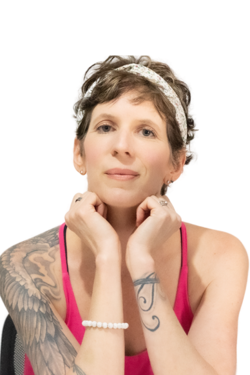 A person with short hair and a floral headband rests their chin on their hands. They are wearing a pink top, a bracelet, and have tattoos on their arm. The background is plain white.