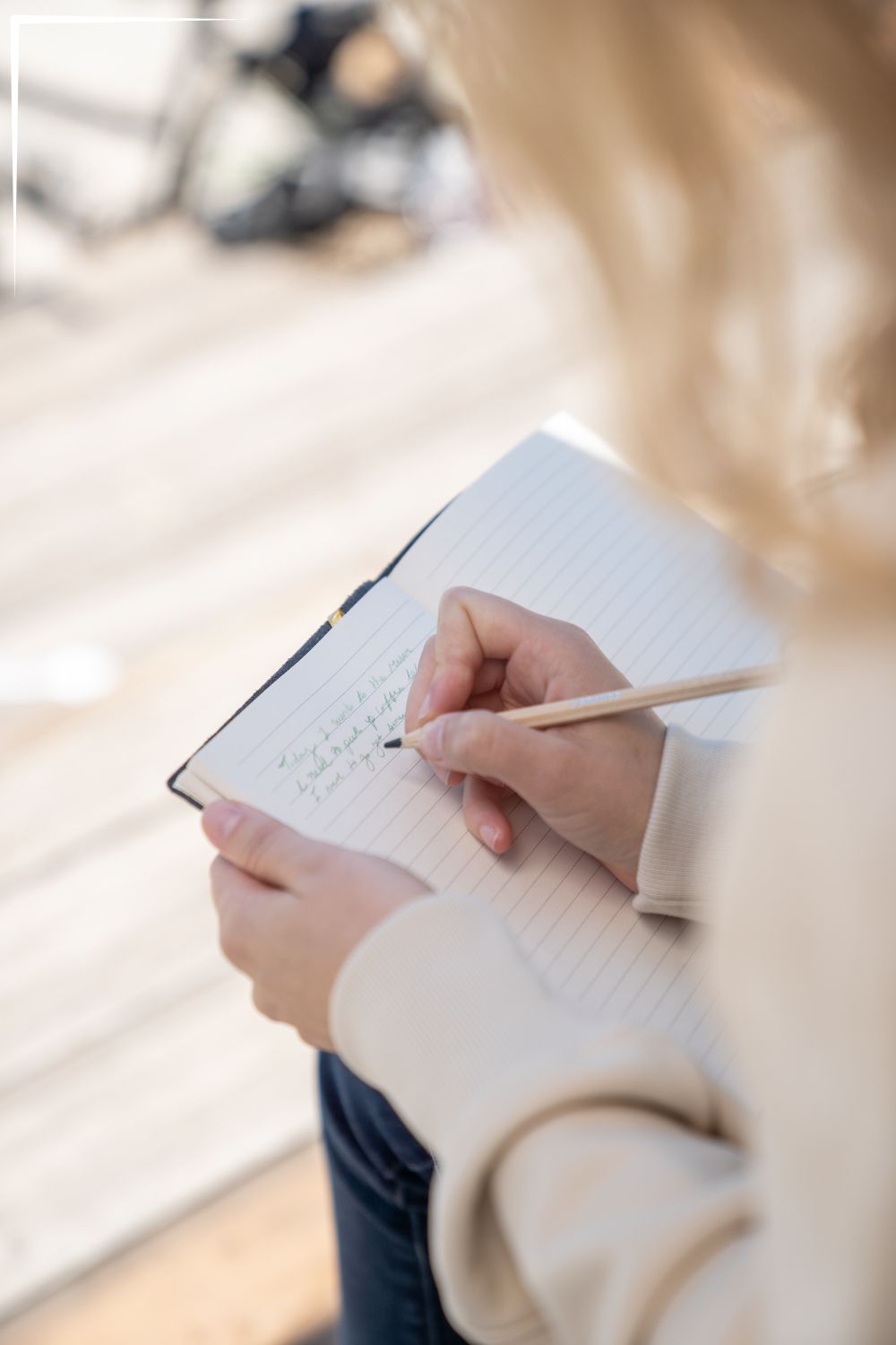 A person writing in a notebook with a pencil. The notebook is held open with one hand, and the page is lined. The background is blurred, focusing on the writing activity.