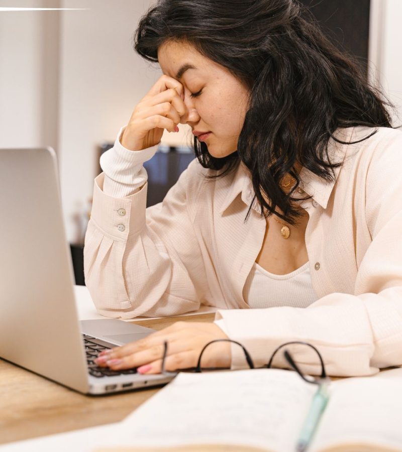 A woman sitting at a table with her hand on her forehead, appearing stressed. She is using a laptop, with glasses and an open notebook on the table, in a well-lit room with a blurred background.