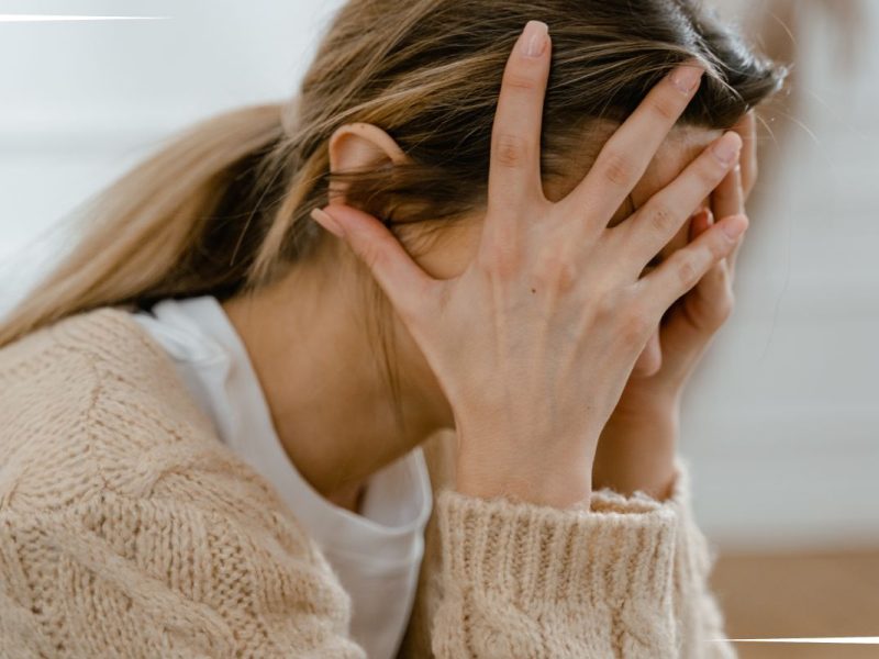 A person wearing a beige sweater and a white shirt sits with hands covering their face, conveying a sense of distress or frustration. The background is softly blurred, focusing attention on the subject.